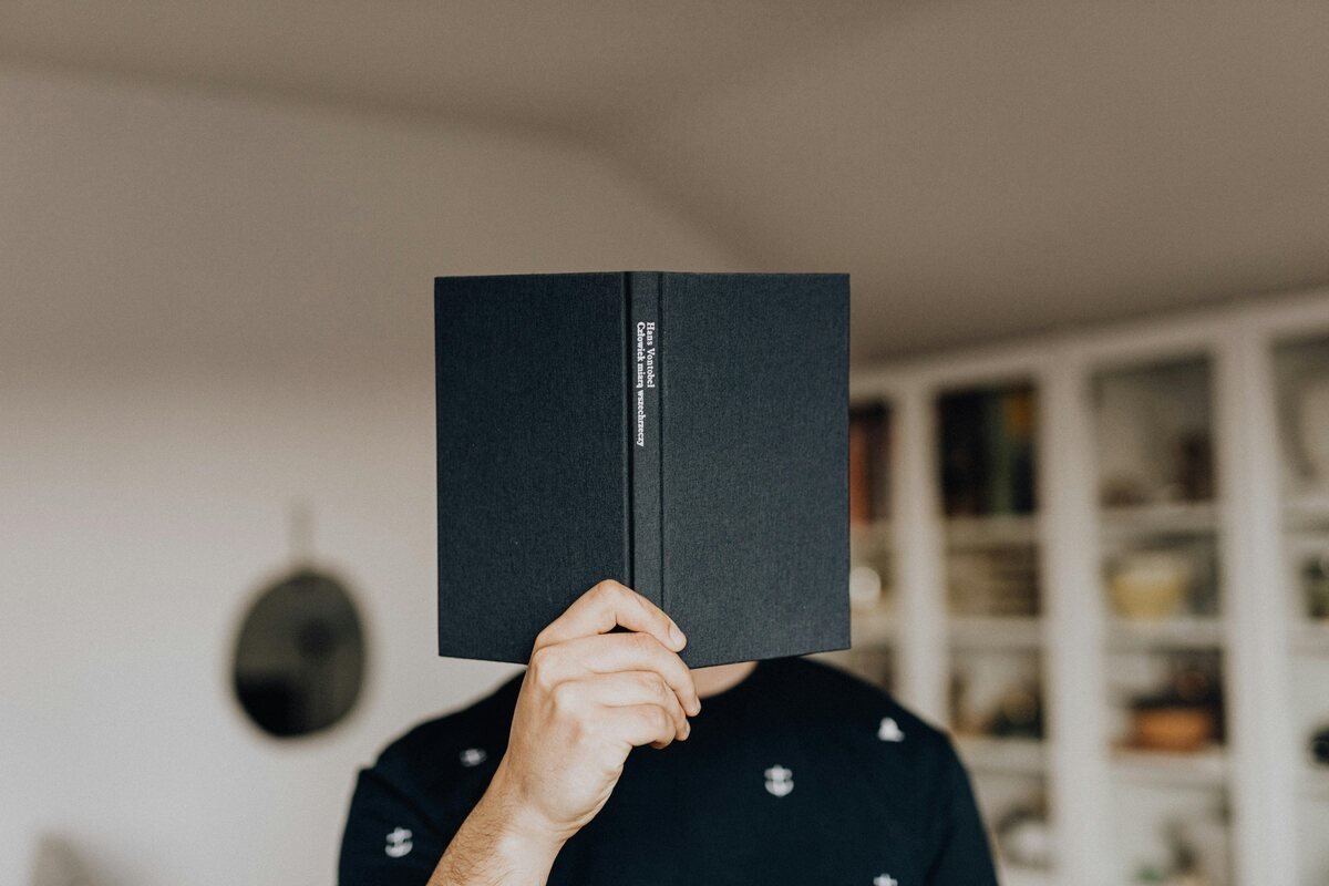 Person holding a black book covering their face, standing in a cozy room with shelves in the background.