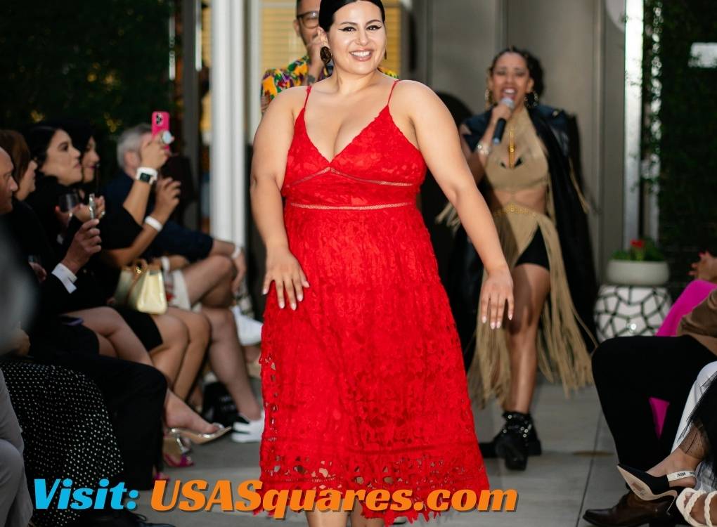 Confident woman in a vibrant red lace dress walking down the runway during a fashion show, with an audience and live music in the background.