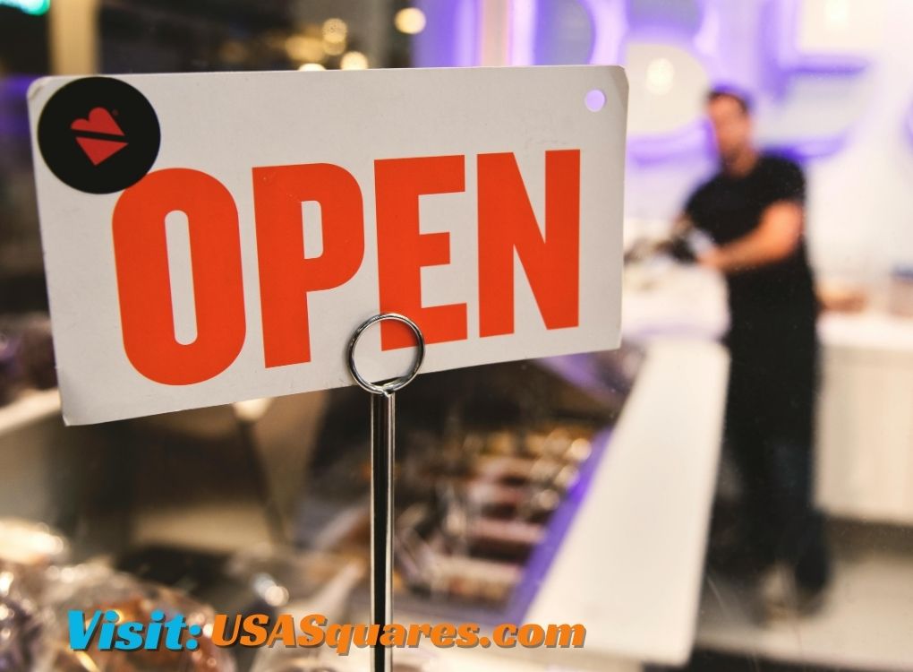 Open sign displayed at a storefront counter with a blurred background of a shopkeeper, emphasizing a welcoming business atmosphere