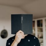 A person holding a black hardcover book in front of their face, obscuring their identity, in a cozy home setting with bookshelves in the background. The book's spine features text, adding intrigue.